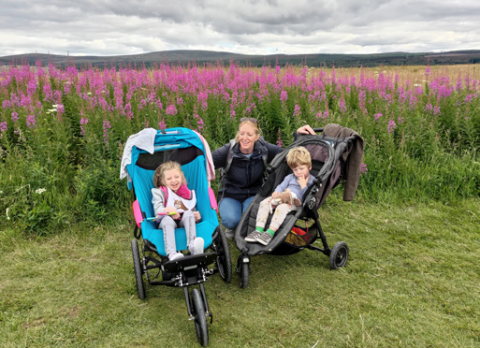 Photo of Laura with Sarah and a young boy who are both in buggies. They are in a meadow with tall pink flowers behind them,