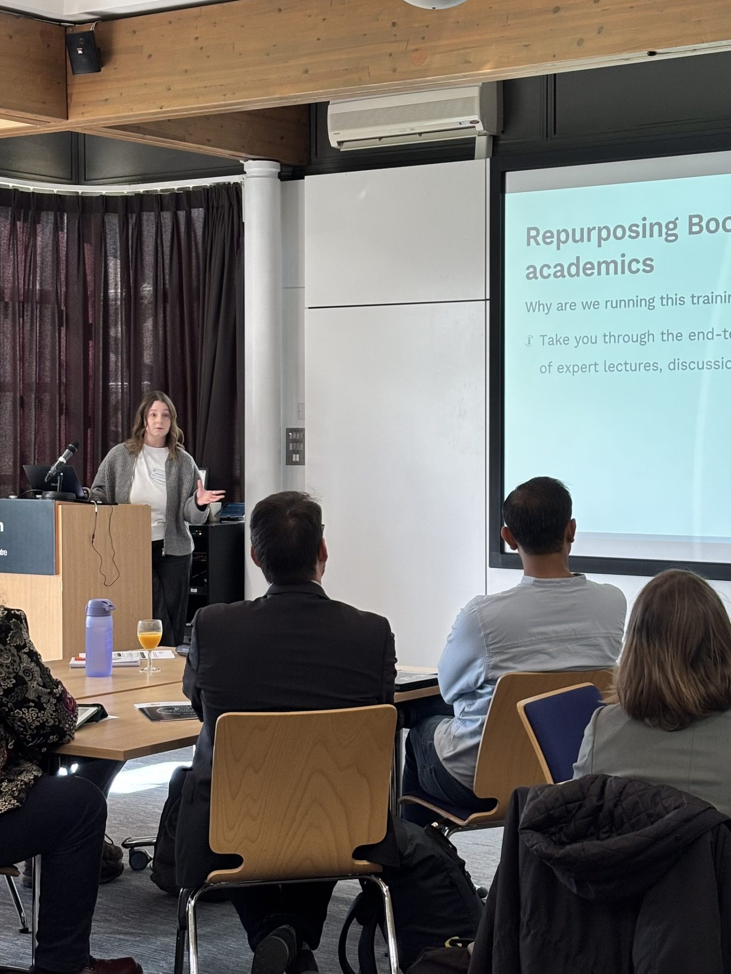 Megan Burley behind a lectern, speaking at the bootcamp with a PowerPoint presentation. Attendees are sat on chairs listening.